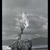 הנפת דגל הדיו באום רשרש, אילת. ב | החייל אברהם אדן (ברן) מטפס על עמוד כדי להניף את דגל הדיו אשר אולתר ע"י חיילי החטיבה, 1949 .