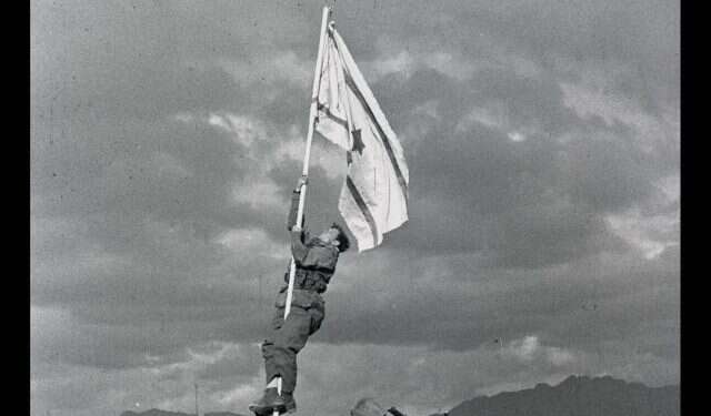 הנפת דגל הדיו באום רשרש, אילת. בצילום, החייל אברהם אדן (ברן) מטפס על עמוד כדי להניף את דגל הדיו אשר אולתר ע בעקבות גלגוליו ההיסטוריים של דגל המדינה